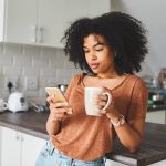 Eine Person steht mit einer Kaffeetasse in der Hand in der Küche und schaut auf ihr Smartphone.
