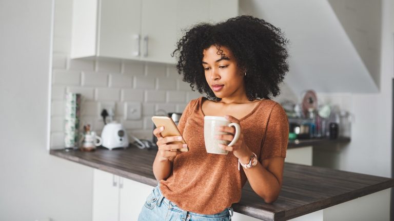 Eine Person steht mit einer Kaffeetasse in der Hand in der Küche und schaut auf ihr Smartphone.