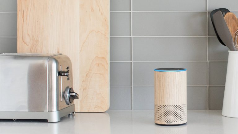 Ein Echo-Lautsprecher im Holz-Look steht auf einer Arbeitsplatte in der Küche neben einem Toaster und einigen anderen Küchenutensilien.