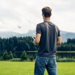 Eine Person steht auf einer Wiese und lässt eine Drohne steigen, die am Horizont verschwommen über einer Baumreihe fliegt.