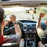 Zwei Personen sitzen in einem Auto und scheinen sich zu Musik zu bewegen. An der Mittelkonsole ist ein Smartphone angesteckt.