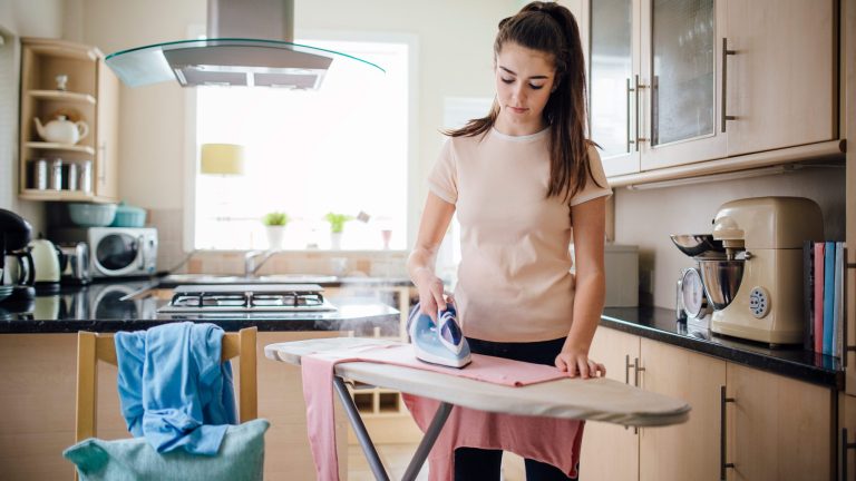 Eine Person steht in einer Küche und bügelt ein rosafarbenes Oberteil.