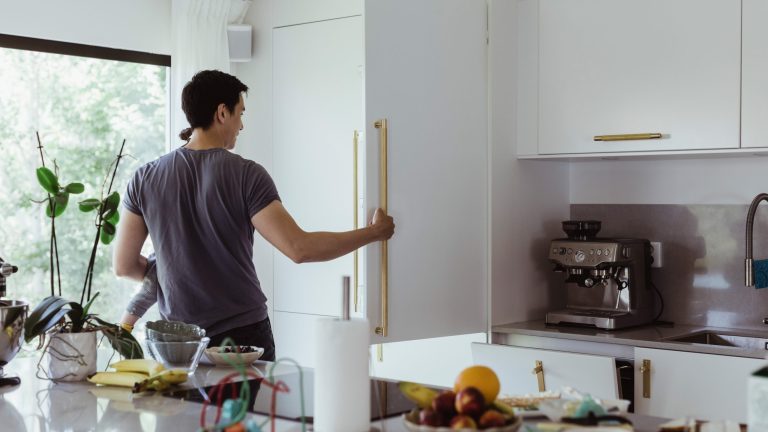 Eine Person mit einem Kind auf dem Arm öffnet den Einbaukühlschrank in einer weißen Küche.