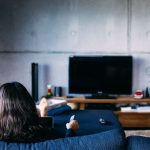 Eine Person sitzt auf einem Sofa vor einem Fernseher und h&auml;lt eine Fernbedienung in der Hand.