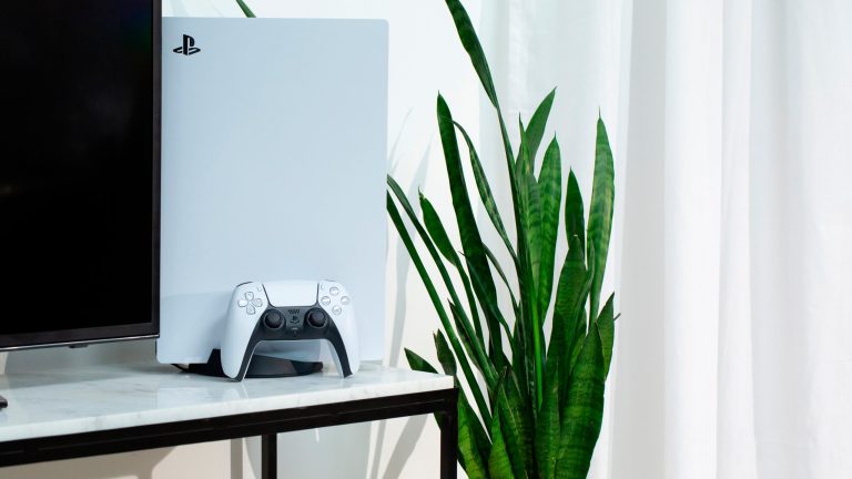Eine PS5 steht neben einem Fernseher auf einem Marmortisch. Vor der Konsole lehnt der DualSense Controller. Neben dem Tisch ragt eine Pflanze in die Höhe.