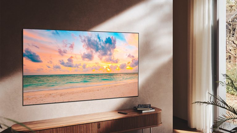 Ein QN 90B hängt an einer Wand. Darunter ist eine TV-Bank zu sehen, auf der ein paar Bücher liegen. Auf dem Display ist ein Strand bei Sonnenuntergang dargestellt.