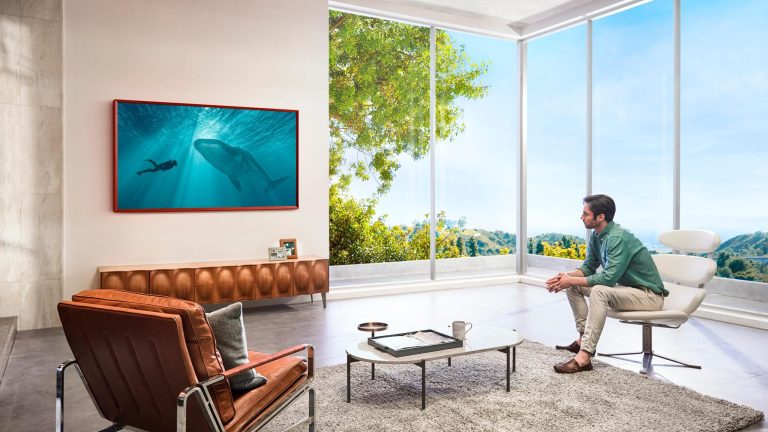 Ein Samsung The Frame hängt an einer Wand in einem Wohnzimmer. Eine Person sitzt auf einem Sessel dafür und blickt auf eine Unterwasser-Szene auf dem Bildschirm.