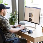 Eine Person sitzt vor einem Bildschirm, auf dem ein Text zu sehen ist. Sie tippt auf einer ergonomischen Tastatur, neben der eine ergonomische Maus steht.