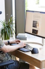 Eine Person sitzt vor einem Bildschirm, auf dem ein Text zu sehen ist. Sie tippt auf einer ergonomischen Tastatur, neben der eine ergonomische Maus steht.