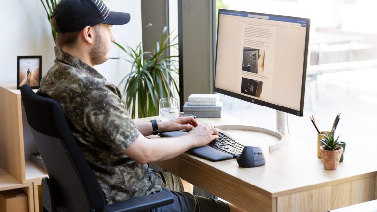 Eine Person sitzt vor einem Bildschirm, auf dem ein Text zu sehen ist. Sie tippt auf einer ergonomischen Tastatur, neben der eine ergonomische Maus steht.