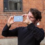Eine Person schießt mit einem iPhone 13 Pro Max ein Foto. Im Hintergrund ist ein Backsteingebäude zu erkennen.