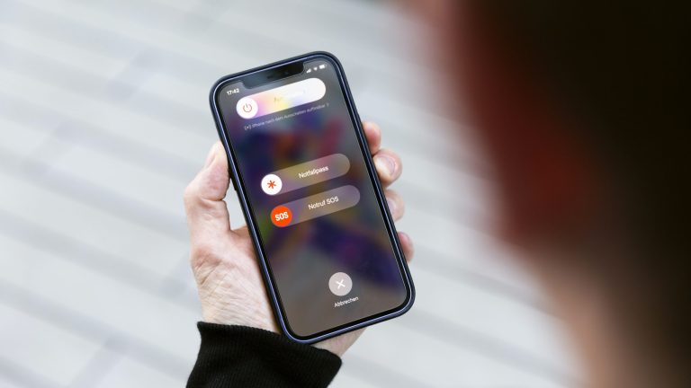 Eine Person hält ein iPhone 12 in der Hand und betätigt die Seitentaste sowie eine der Lautstärke-Tasten. Auf dem Display sind die Optionen zu sehen, dass Gerät auszuschalten, ein, den Notfallpass zu aktivieren oder einen Notruf abzusetzen.