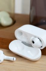 AirPods Pro Ein Paar AirPods Pro liegt samt Ladecase auf einem Holzbrett. Einer der beiden Kopfhörer liegt dabei vor der geöffneten Hülle.