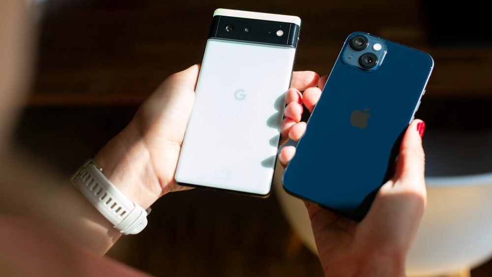 Zwei Hände halten jeweils ein Smartphone: Einmal ein weißes Google Phone und einmal ein dunkelblaues iPhone.