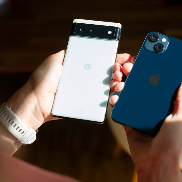 Zwei H&auml;nde halten jeweils ein Smartphone: Einmal ein wei&szlig;es Google Phone und einmal ein dunkelblaues iPhone.