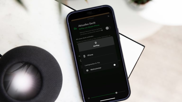 Ein schwarzer HomePod steht auf einem Tisch. Daneben liegt ein Smartphone, auf dem die Spotify-App geöffnet wurde.