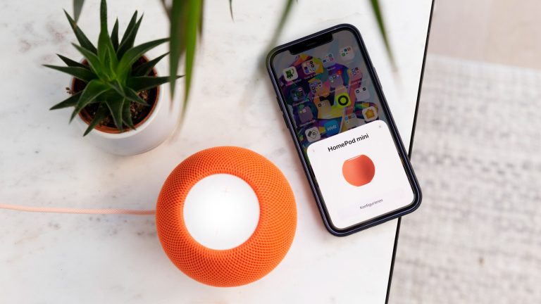Ein iPhone liegt neben einem HomePod mini in Orange. Auf dem Display des Handys ist der erkannte HomePod zu sehen.