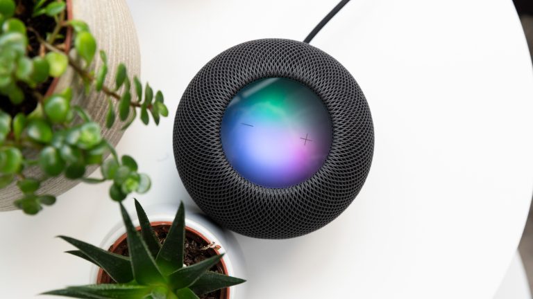 Ein schwarzer HomePod mini steht auf einem weißen Tisch. Auf dem Display des Lautsprechers sind wabernde Farben zu erkennen, die signalisieren, dass Siri aktiv ist.