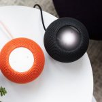 Zwei HomePods mini in Schwarz und Orange stehen auf einem weißen Tisch nebeneinander.