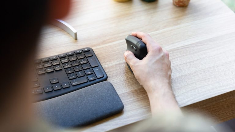 Eine Person sitzt an einem Schreibtisch und h&auml;lt eine ergonomische Maus in ihrer rechten Hand. Daneben ist eine Tastatur mit Handballenauflage zu sehen.