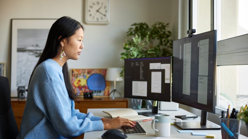 Eine Person sitzt vor zwei Monitoren auf einem Schreibtisch und tippt auf einer flachen Office-Tastatur.