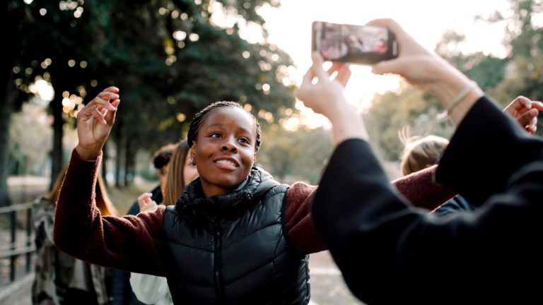 Eine junge Frau tanzt auf offener Straße und lächelt in eine Smartphone-Kamera. Im Vordergrund sieht man Arme, die das Smartphone waagerecht in die Luft halten.