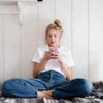 Eine Person sitzt im Schneidersitz auf einem Bett und schaut auf ein Smartphone, das sie in den H&auml;nden h&auml;lt.