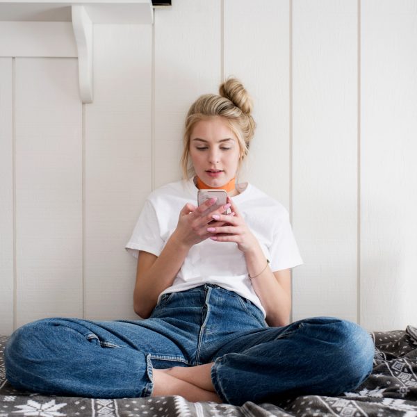 Eine Person sitzt im Schneidersitz auf einem Bett und schaut auf ein Smartphone, das sie in den Händen hält.