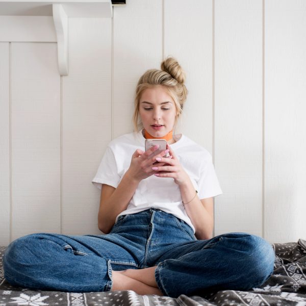 Eine Person sitzt im Schneidersitz auf einem Bett und schaut auf ein Smartphone, das sie in den H&auml;nden h&auml;lt.