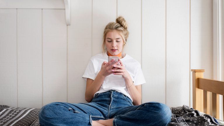 Eine Person sitzt im Schneidersitz auf einem Bett und schaut auf ein Smartphone, das sie in den Händen hält.