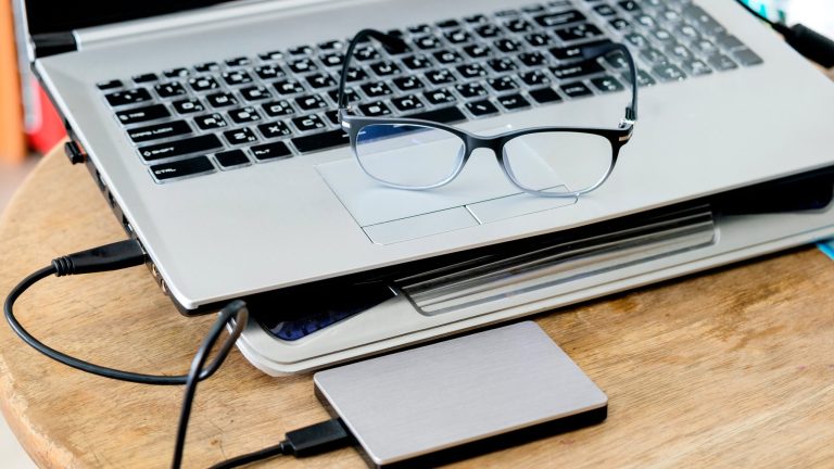 Auf einem Tisch steht ein Laptop mit externer Festplatte, auf ihm liegt eine Brille.