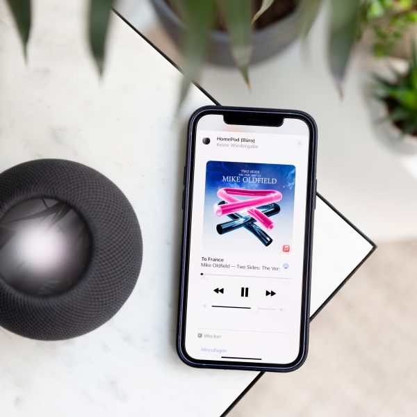 Ein schwarzer HomePod mini steht auf einem Marmortisch. Neben dem Lautsprecher liegt ein iPhone, auf dem Apple Music ge&ouml;ffnet ist.