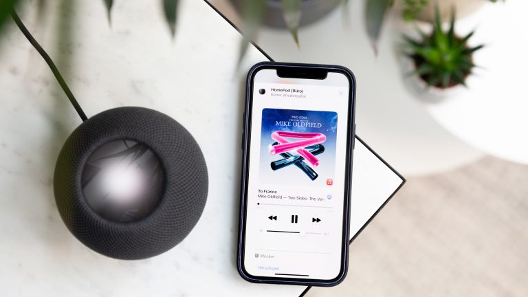 Ein schwarzer HomePod mini steht auf einem Marmortisch. Neben dem Lautsprecher liegt ein iPhone, auf dem Apple Music geöffnet ist.