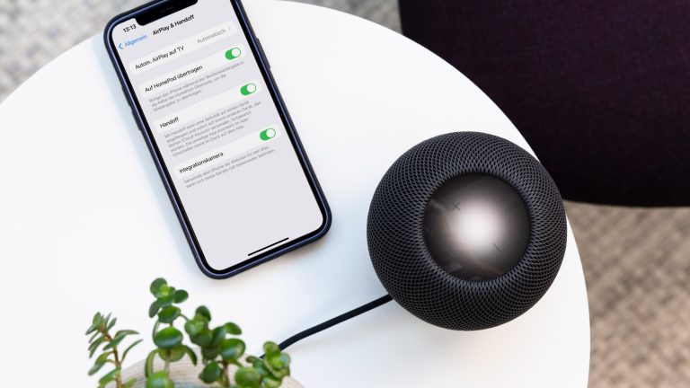 Auf einem weißen, runden Tisch steht einer schwarzer HomePod mini. Daneben liegt ein iPhone, auf dem gerade die Einstellungsoptionen zu Handoff angezeigt werden.