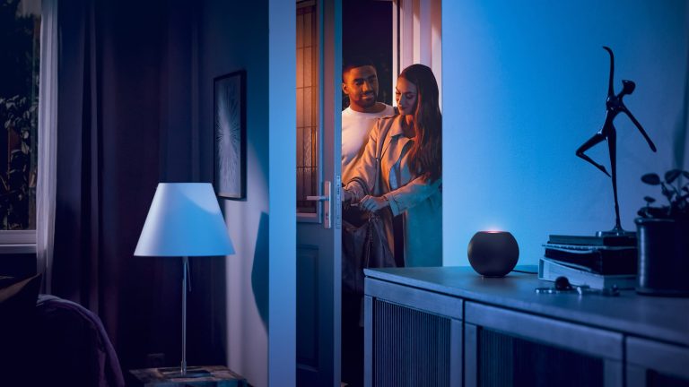 Zwei Personen betreten eine Wohnung, in der auf einer Anrichte ein HomePod mini in Schwarz steht.