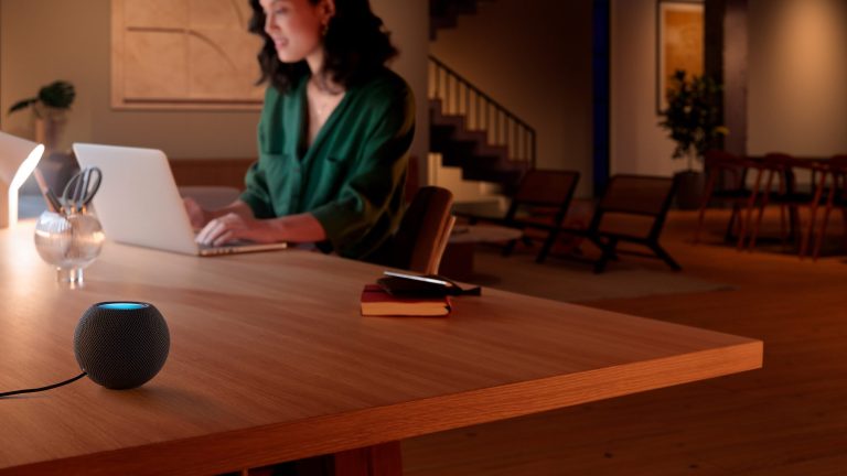 Eine Person sitzt an einem Tisch vor einem MacBook. Im Vordergrund steht auf dem Tisch eine HomePod mini.