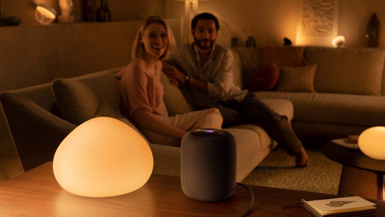 Zwei Personen sitzen auf einer Sofalandschaft und blicken in Richtung eines HomePods, der im Vordergrund neben einer Hue-Leuchte steht.
