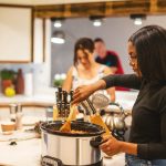 Eine Person steht vor einem Slow-Cooker-Kochtopf und r&uuml;hrt darin mit einem Holzl&ouml;ffel.