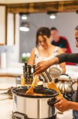Eine Person steht vor einem Slow-Cooker-Kochtopf und rührt darin mit einem Holzlöffel.