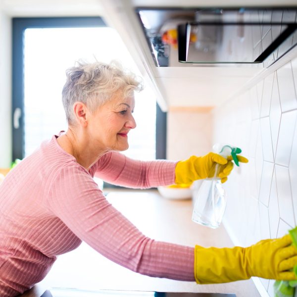 Eine Person putzt die Fliesen ihrer Küchenzeile. Hierbei benutzt sie gelbe Handschuhe und eine Sprühflasche.