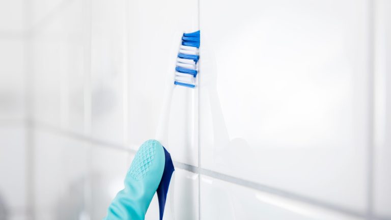 Eine Hand führt eine blau-weiße Zahnbürste eine Fliesenfuge entlang.