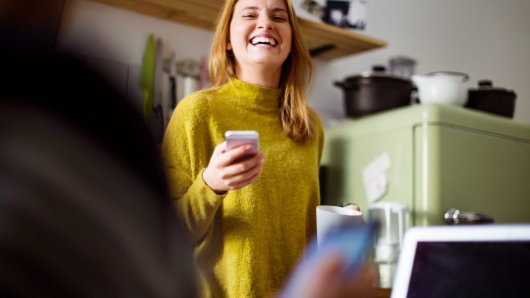 Eine lachende Frau steht in einer K&uuml;che und h&auml;lt ein Smartphone in der Hand.