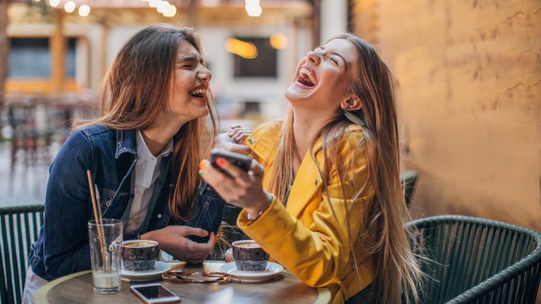 Zwei Personen sitzen in einem Caf&eacute; und lachen ausgelassen. Die eine Person h&auml;lt ein Smartphone in der Hand.