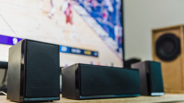 Drei Lautsprecher stehen vor einem Fernseher in einem Raum. Im Hintergrund ist ein Subwoofer zu sehen.