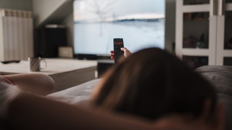 Eine Person liegt auf einem Sofa und hält eine Fernbedienung in die Höhe. Im Hintergrund ist unscharf ein Fernseher zu erkennen, auf dem gerade eine Schneelandschaft gezeigt wird. 