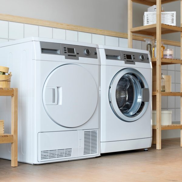 Ein Trockner steht in einem Badezimmer neben einer Waschmaschine. Beide Ger&auml;te sind gerahmt von Holzregalen, auf denen diverse Reinigungsmittel stehen.