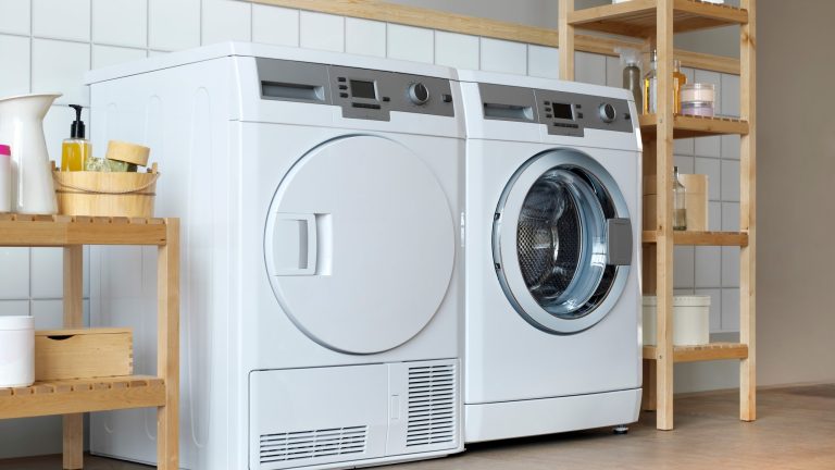 Ein Trockner steht in einem Badezimmer neben einer Waschmaschine. Beide Geräte sind gerahmt von Holzregalen, auf denen diverse Reinigungsmittel stehen.