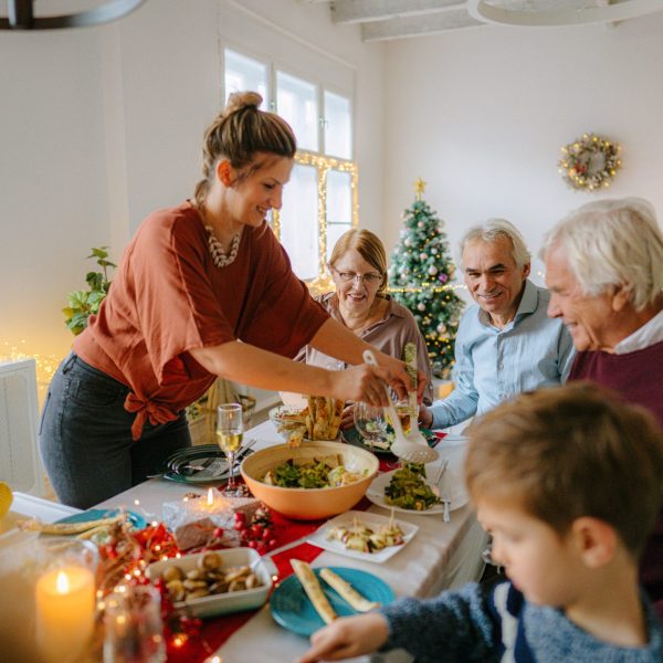 Mehrere Personen sitzen an einem langen Esstisch, auf dem mehrere Schalen mit Speisen stehen. Im Hintergrund leuchtet ein Weihnachtsbaum.