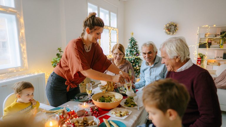 Mehrere Personen sitzen an einem langen Esstisch, auf dem mehrere Schalen mit Speisen stehen. Im Hintergrund leuchtet ein Weihnachtsbaum.