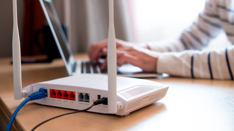 Blick auf die Rückseite eines weißen WLAN-Routers, in den zwei Kabel führen. Dahinter sitzt eine Person und tippt auf einem Laptop.